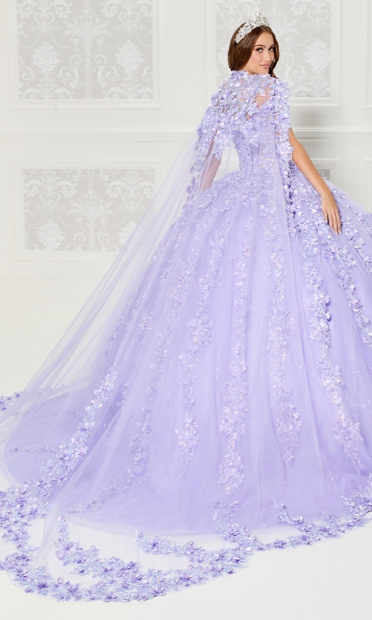 A woman in the Princesa PR30120 lavender floral tulle quinceanera dress with 3D flowers and a matching long cape stands in an elegant white room.
